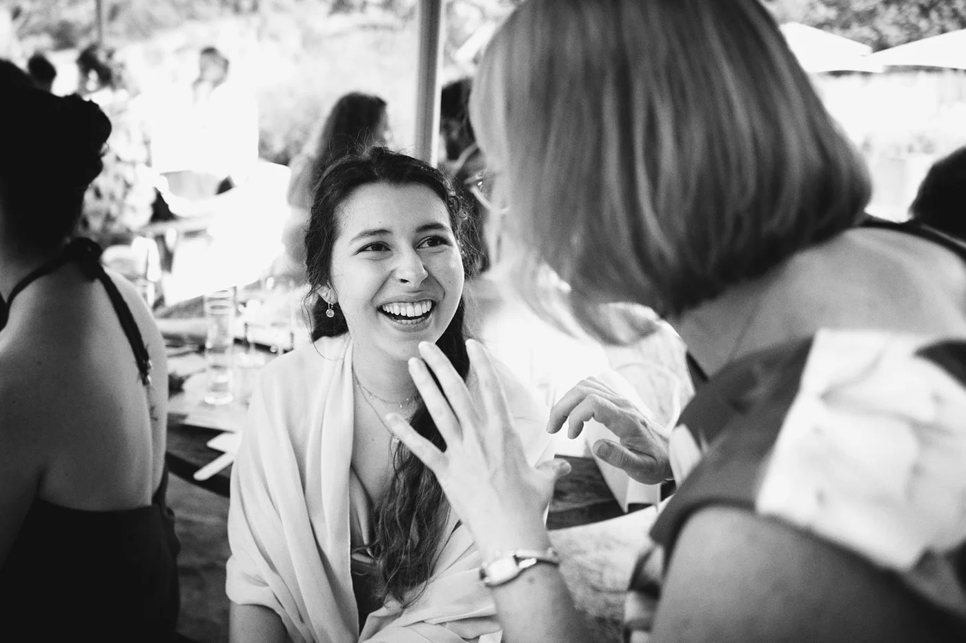 A young woman with long, dark hair and earrings is smiling and laughing while talking to an older woman with blond hair at a social gathering.