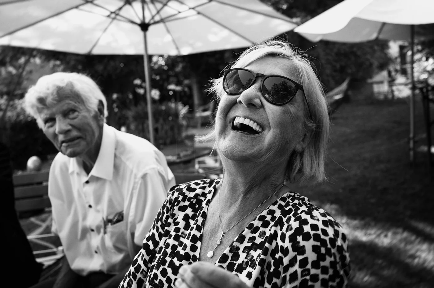 Black and white photo of a woman laughing wearing sunglasses and a leopard print dress, with an older man with white hair sitting in the background outdoors under umbrellas.