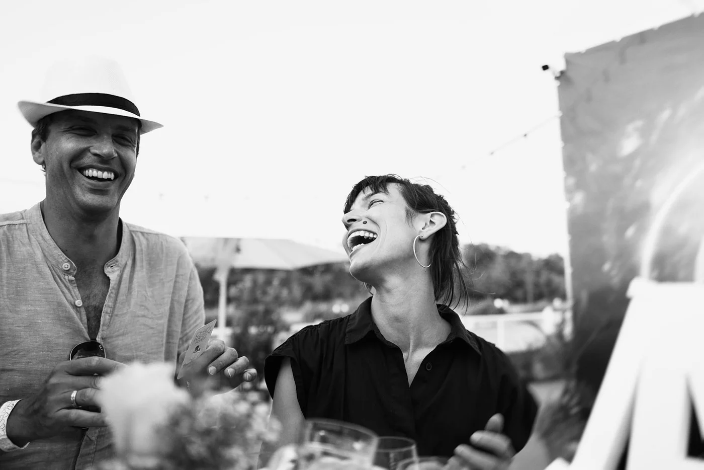 Two people laughing and enjoying themselves at an outdoor event, with one man wearing a hat and holding a card, and a woman wearing hoop earrings.