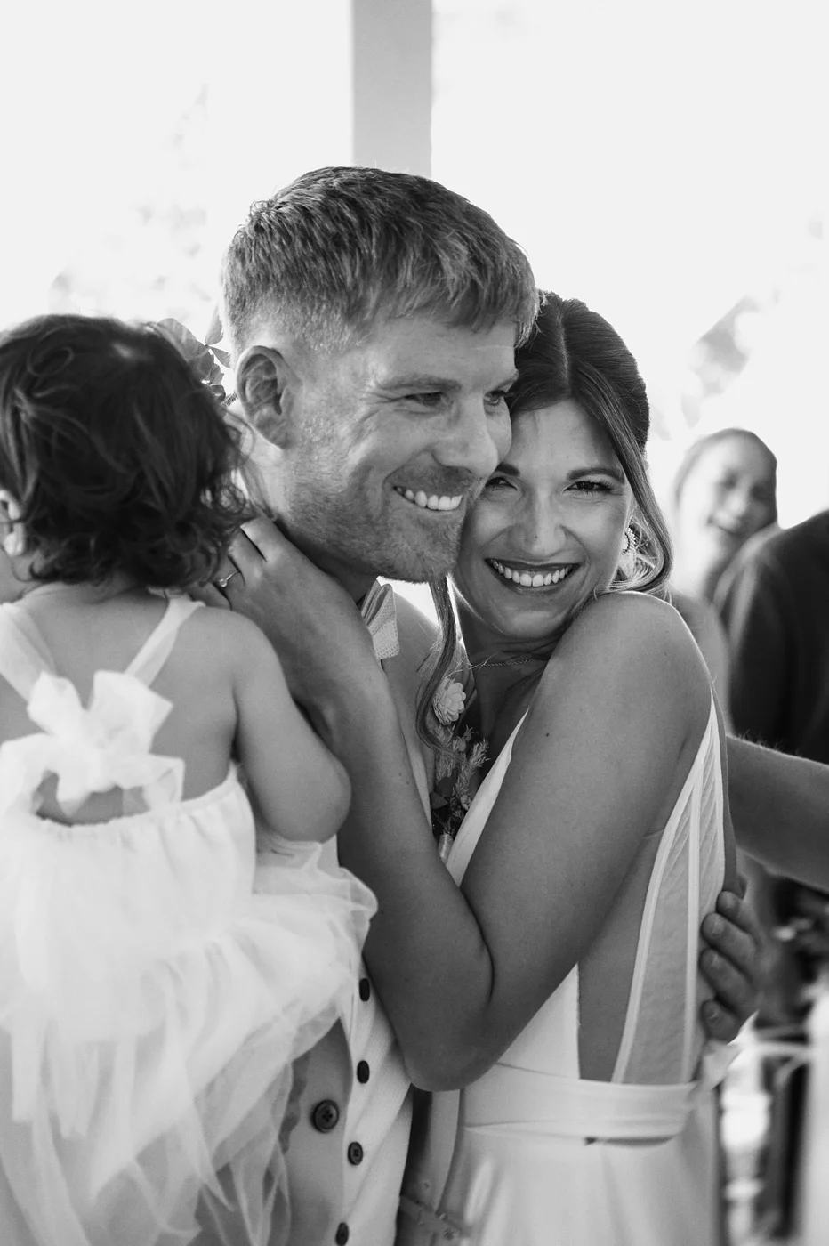 A black and white photo of a joyful couple embracing and smiling, holding a young girl with curly hair in a sleeveless dress.