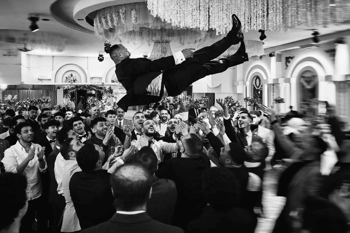 A black and white photo of a wedding celebration with a groom being tossed into the air by guests in a decorated banquet hall.