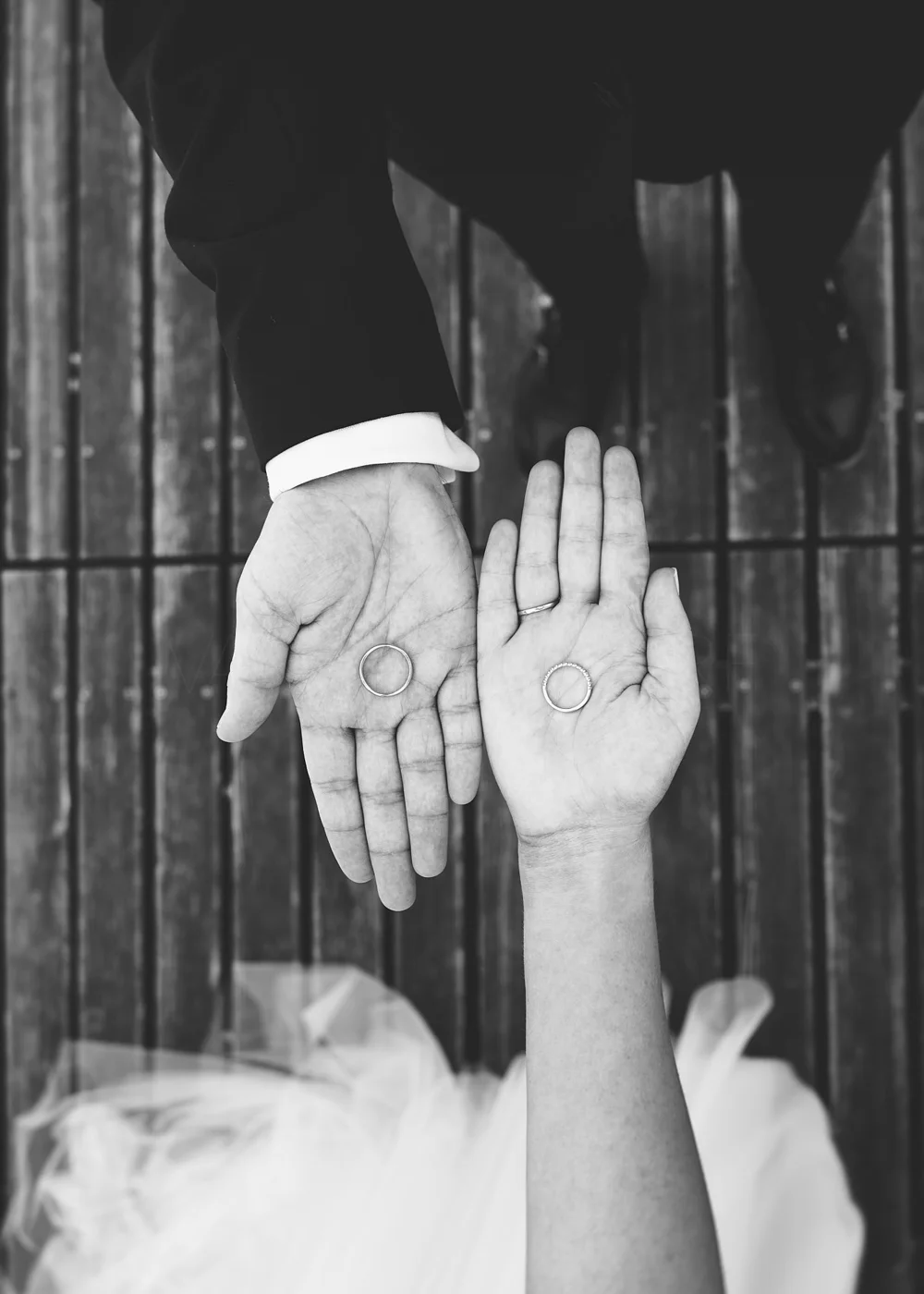 Black and white photo of a couple holding wedding rings over a wooden floor, viewed from above.