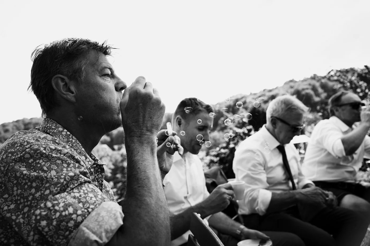 Four men sit outdoors, one blowing bubbles, all wearing light shirts and sunglasses. The photo is in black and white.