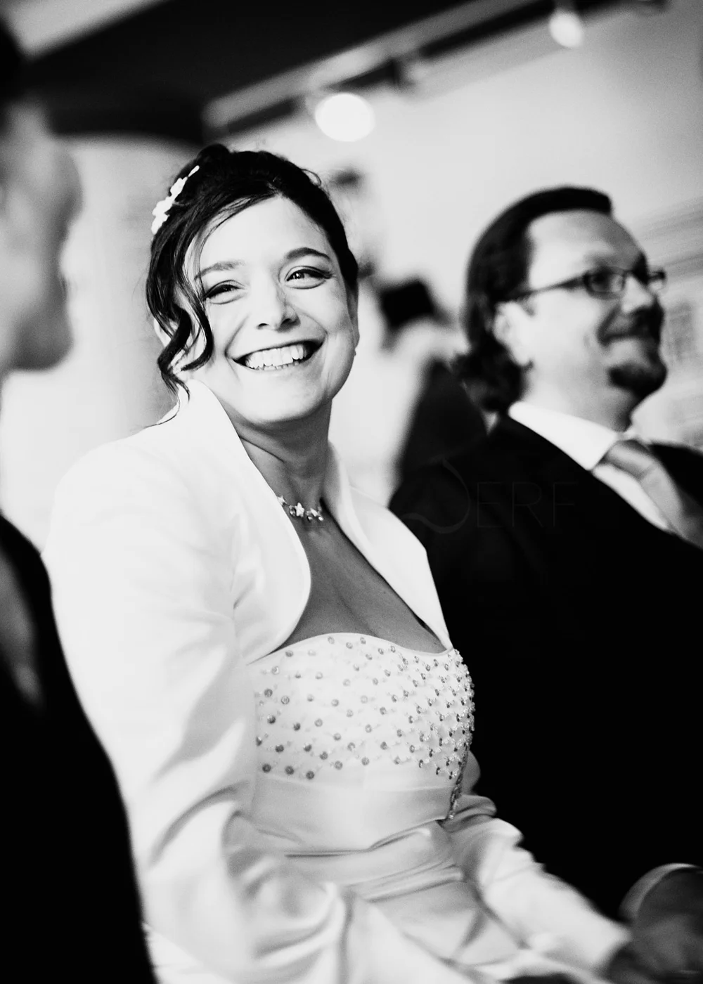 Black and white photo of a woman in wedding attire smiling, sitting between two men in formal suits at a social event.