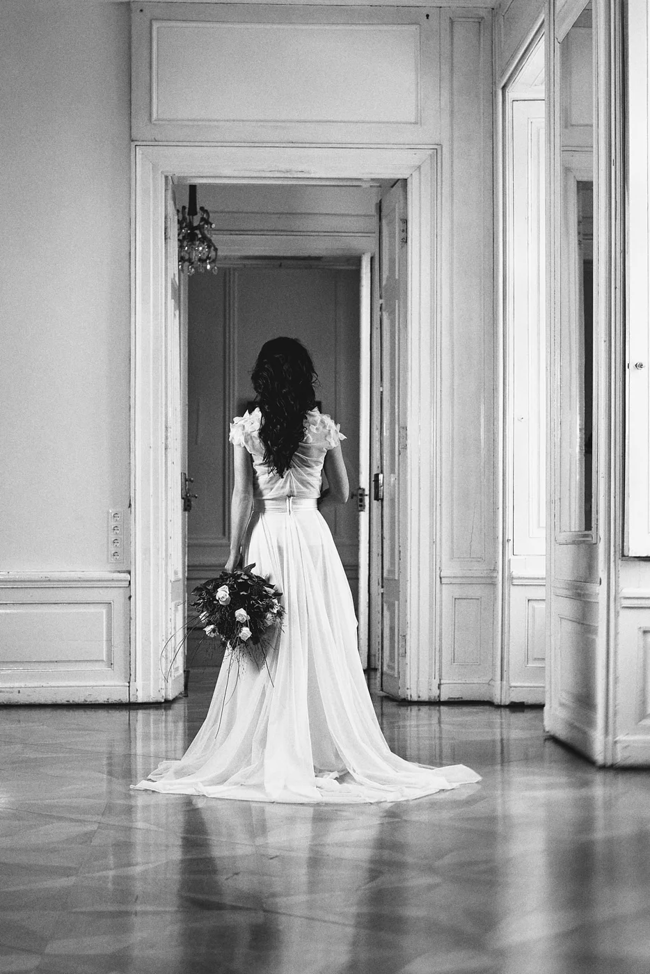 A woman in a white wedding dress holding a bouquet of flowers stands in a room with wooden floors and ornate paneling, facing away from the camera towards an open doorway.