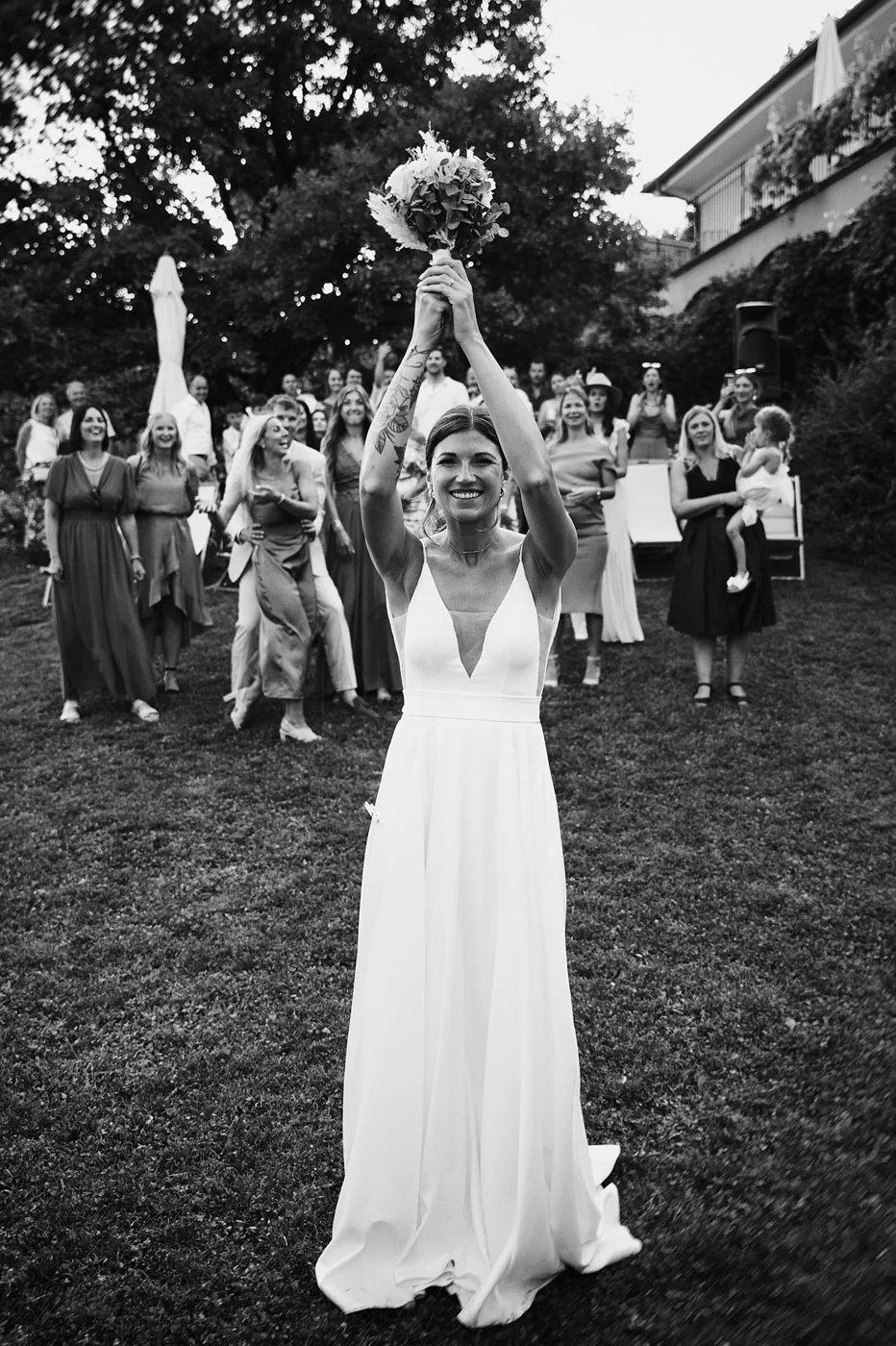 Black and white photo of a smiling bride in a white wedding dress holding a bouquet above her head, standing on grass with a group of women in dresses and a child in the background at an outdoor wedding celebration.