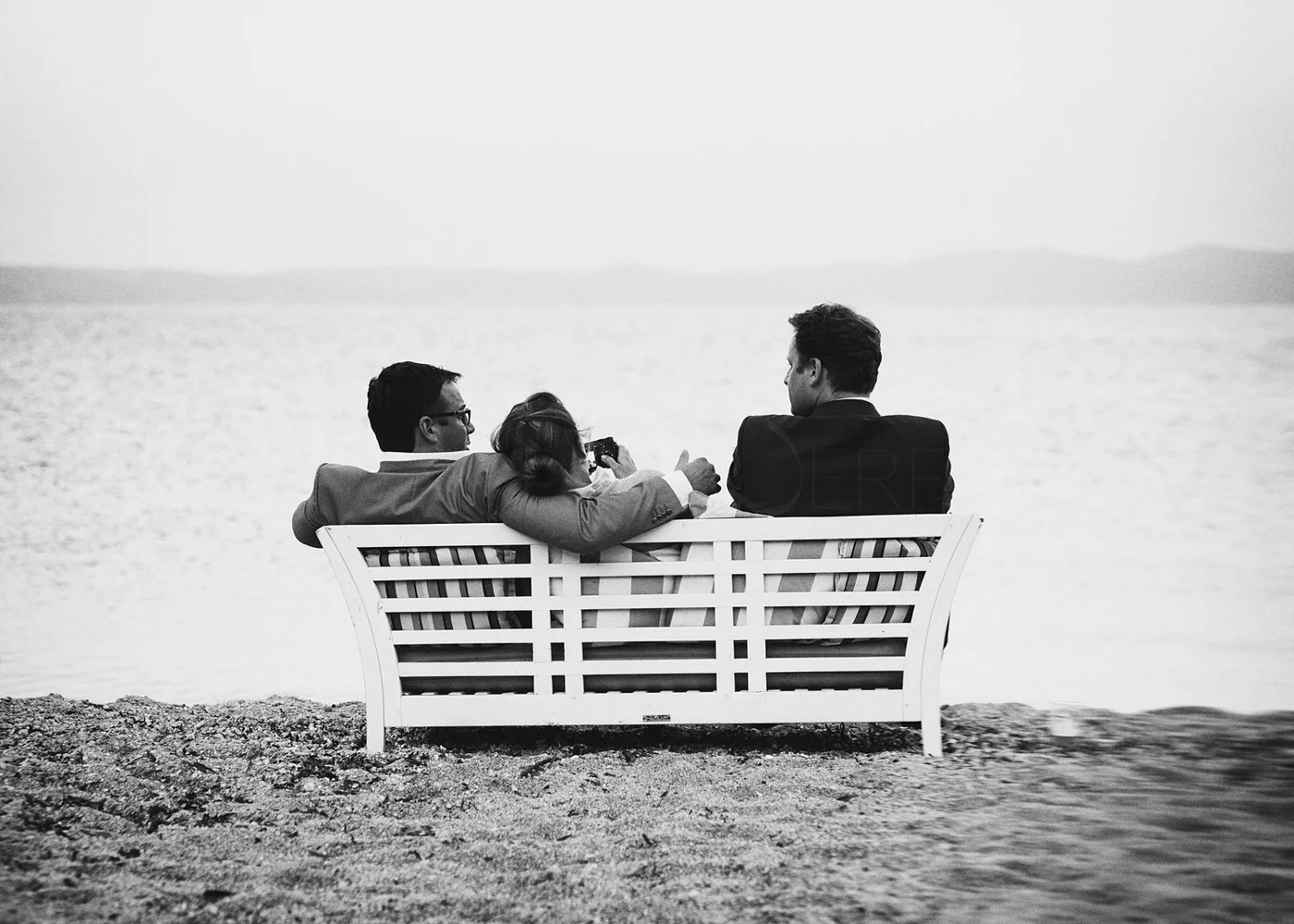 Three people in formal attire sitting on a white bench by water, talking and taking photos, black and white photo.