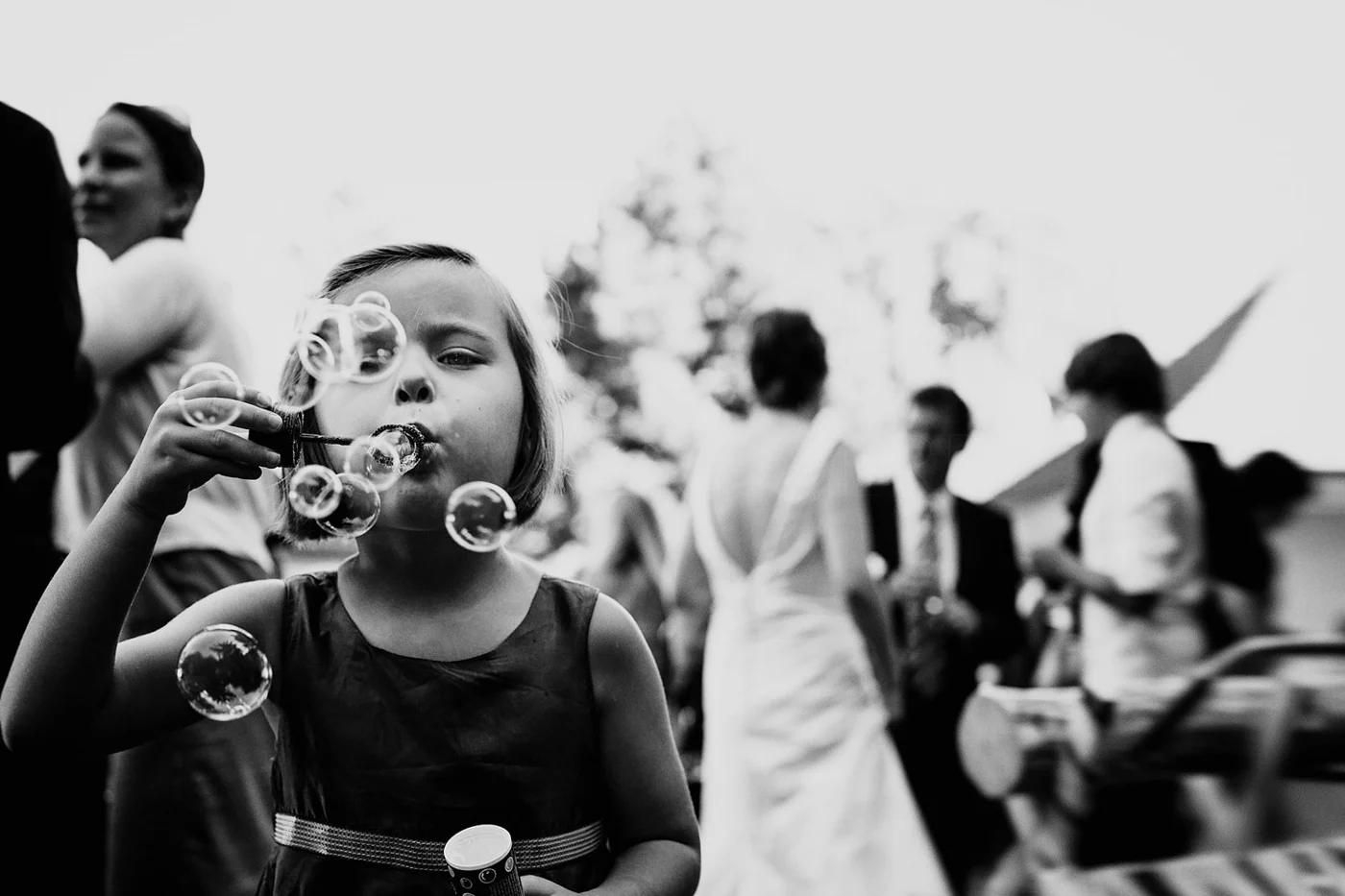 A young girl blows bubbles at an outdoor gathering while several people stand and sit in the background.