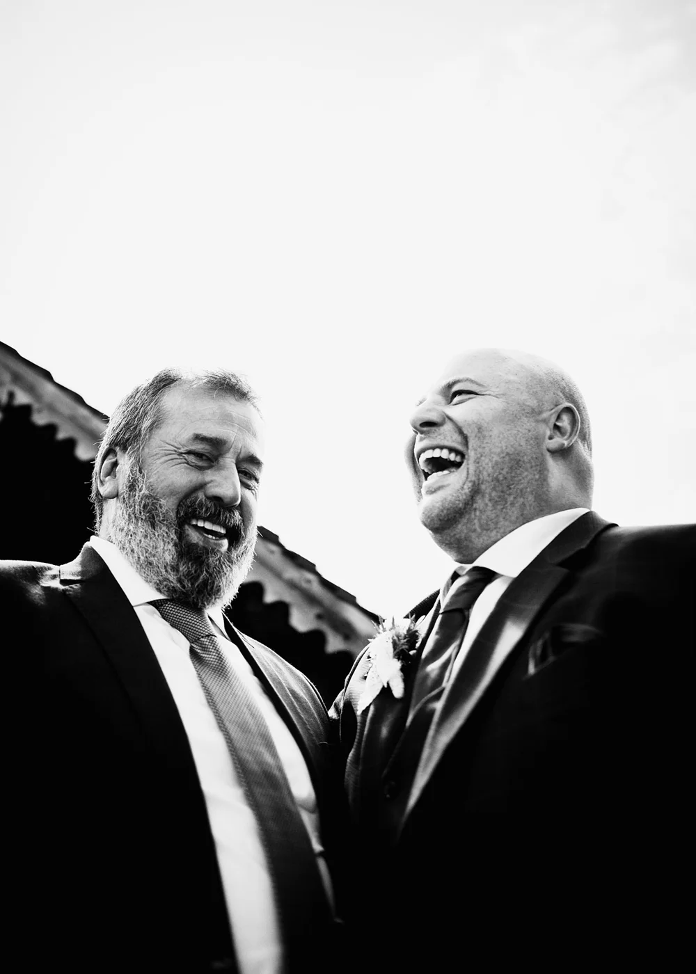 Two men in suits laughing and smiling at an outdoor event, taken from a low angle in black and white.