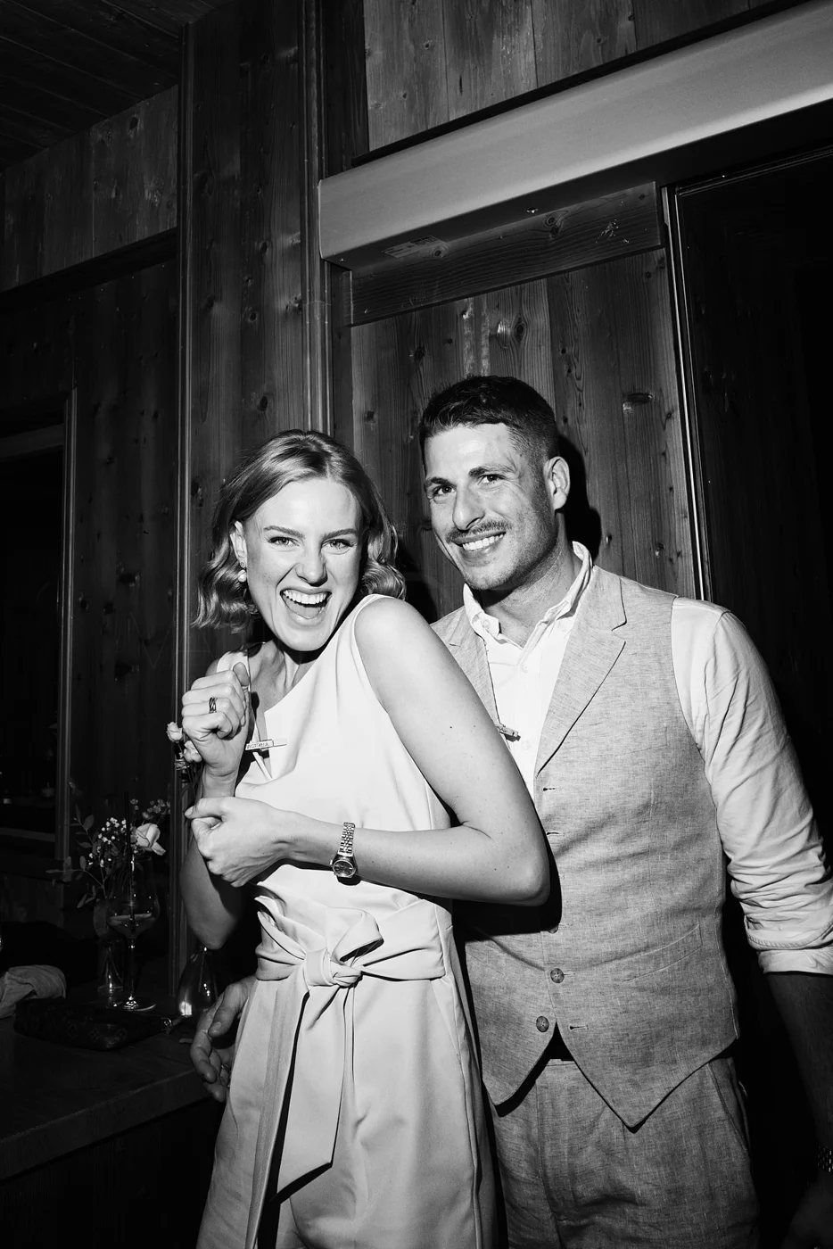 Black and white photo of smiling woman and man standing close together, celebrating, indoors with wooden wall background.