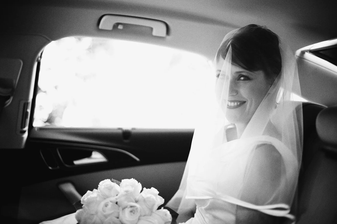 Black and white photo of a smiling bride sitting in the back of a car, wearing a wedding dress and veil, holding a bouquet of white roses.