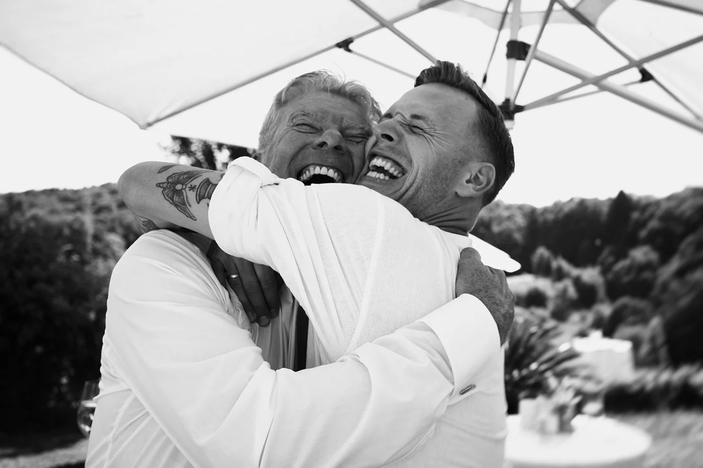 Two men embrace and laugh together outdoors under a canopy, sharing a joyful hug.