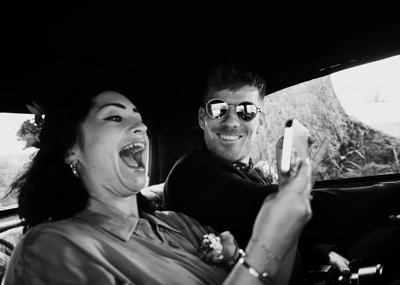 A woman and a man riding in a vehicle, smiling as she looks at her phone. The woman appears excited with her mouth open, and the man, wearing sunglasses, is smiling at her.