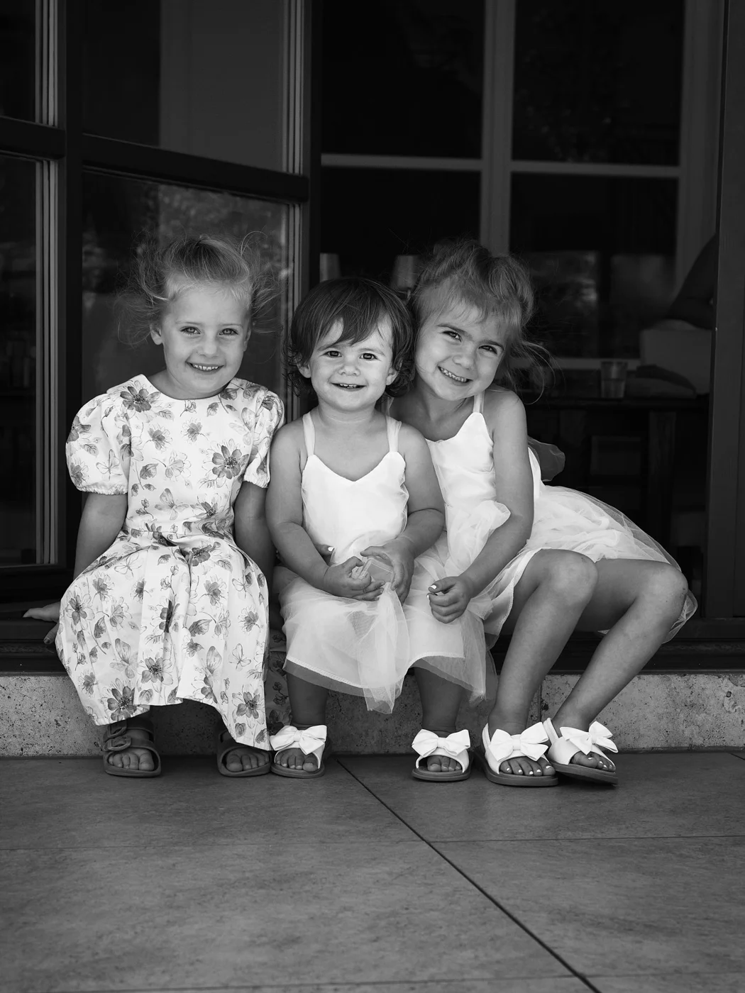 Black and white photo of three young girls sitting on a porch, smiling at the camera. They are dressed in light-colored dresses and sandals.