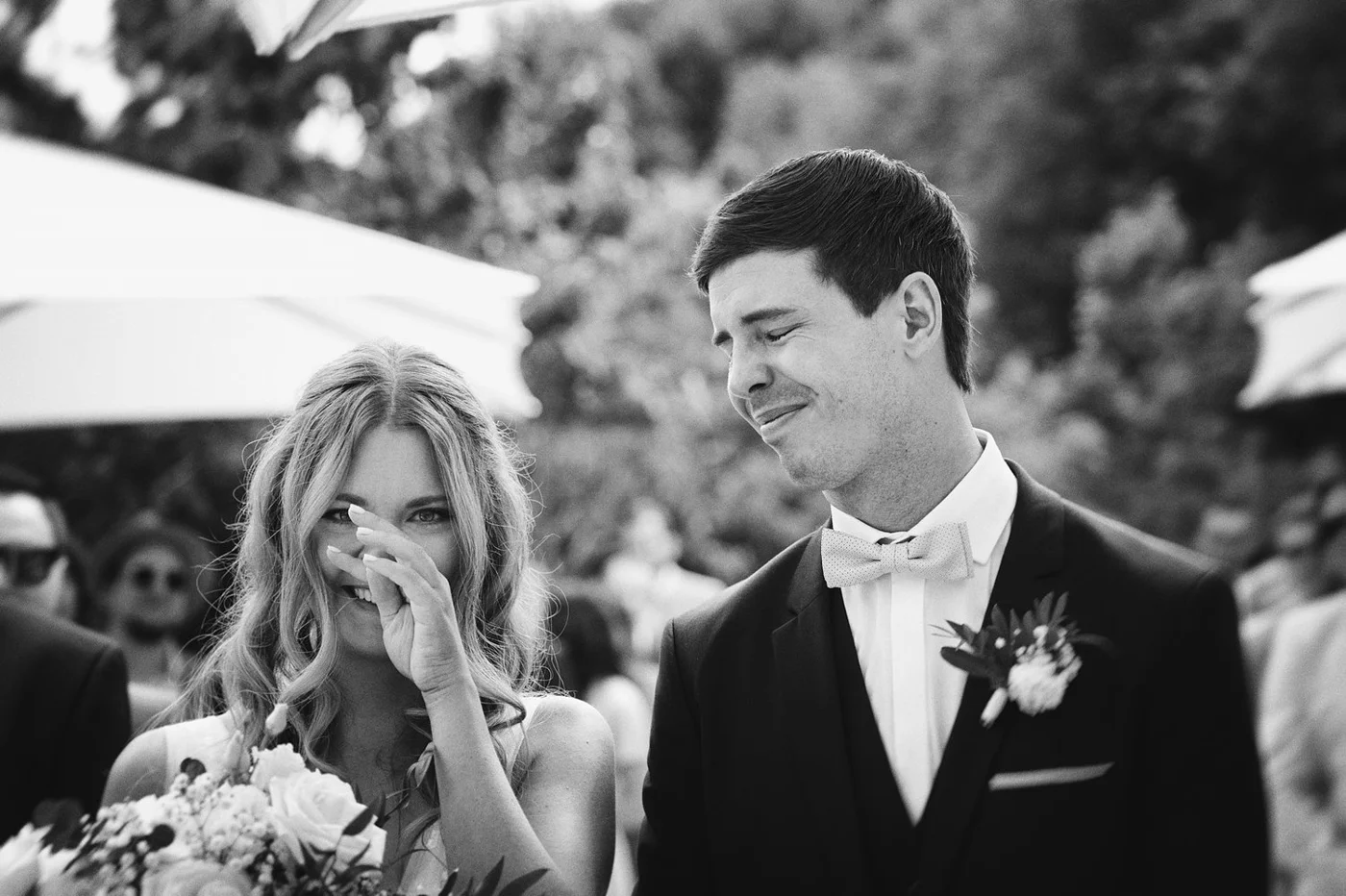 A bride and groom at an outdoor wedding, with the bride smiling and covering her mouth, and the groom smiling with his eyes closed, dressed in a black tuxedo with a bow tie and boutonniere, surrounded by guests and umbrellas.