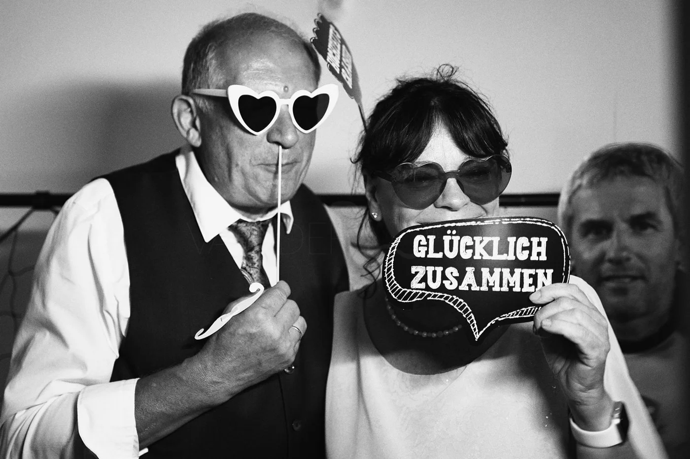 Three people at a festive event wearing sunglasses and holding signs, including one speech bubble with German text. The photo is in black and white.