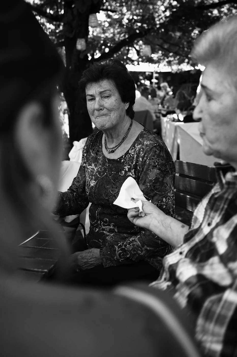 Black and white photo of an elderly woman with short dark hair, wearing a patterned top and a necklace, sitting outdoors at a table with others, under a large tree with dappled sunlight.