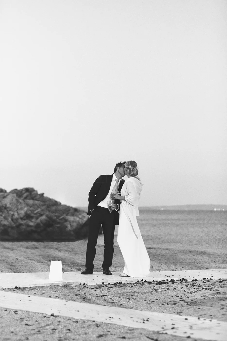 A black and white photo of a couple in wedding attire sharing a kiss on a beach, holding glasses.
