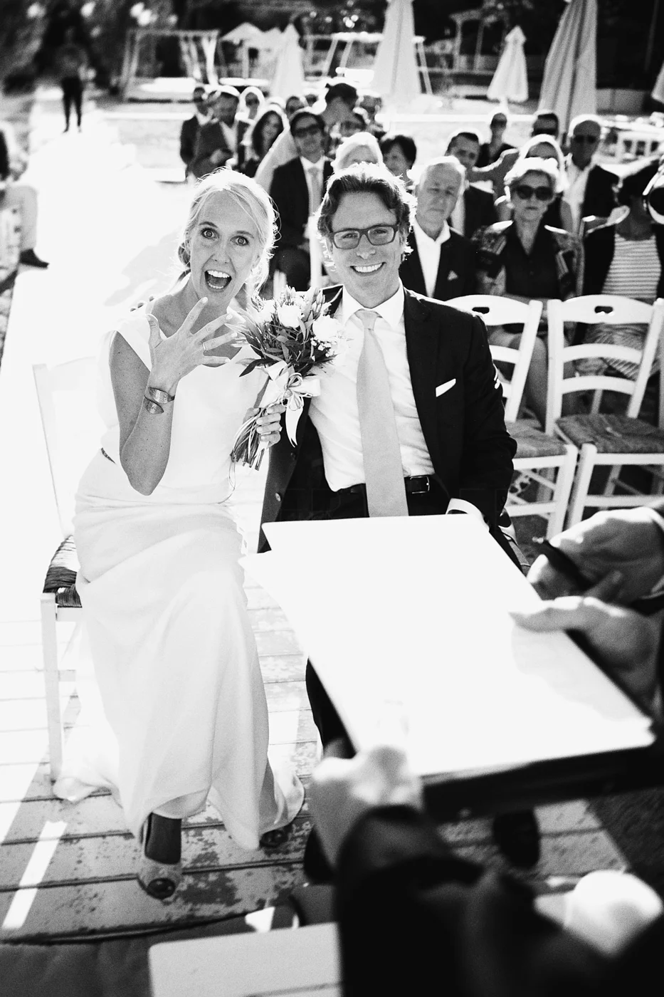 Black and white photo of a wedding ceremony showing a bride and groom seated with an audience behind them. The bride is holding a bouquet and has her hand raised in an excited gesture. The groom is wearing glasses and a suit. The crowd is seated outd