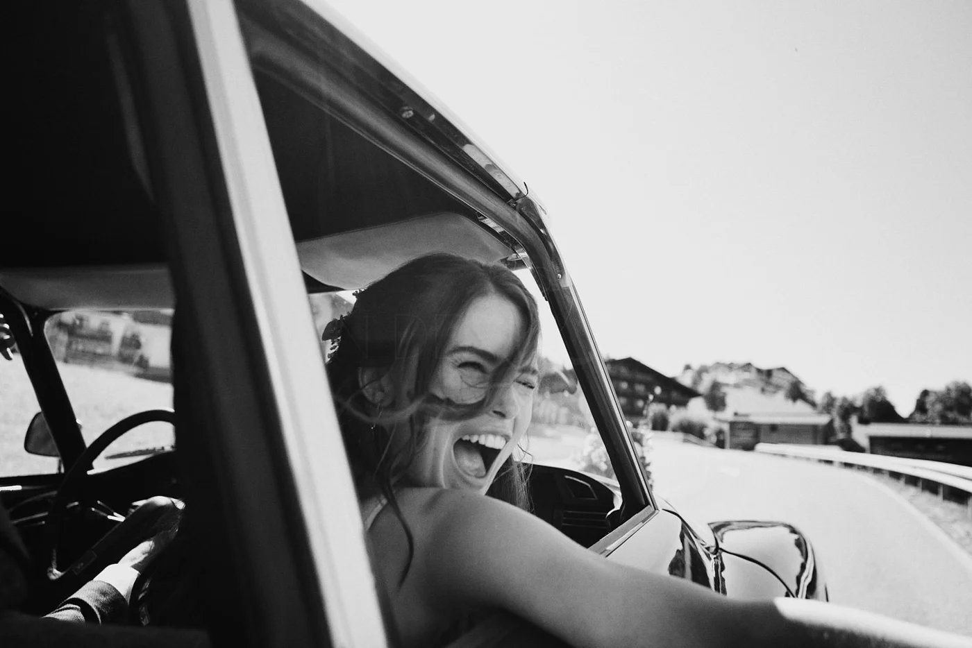 A woman with long hair is smiling and laughing as she leans out of a car window on a sunny day, with houses and trees in the background.