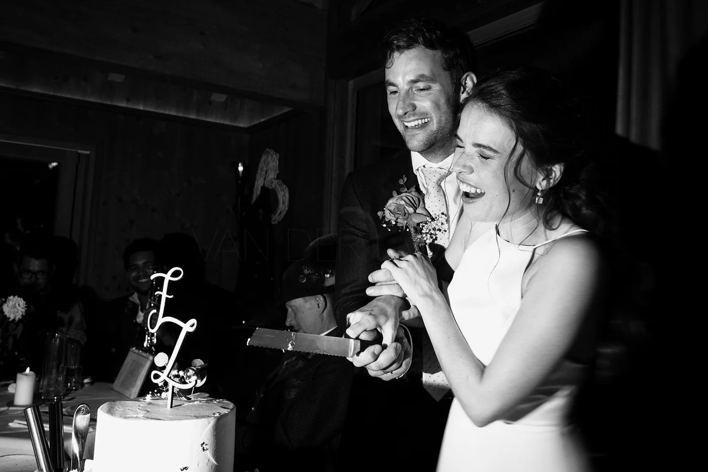 A bride and groom smile and cut a wedding cake together in a joyful moment at their wedding reception, with guests visible in the background.