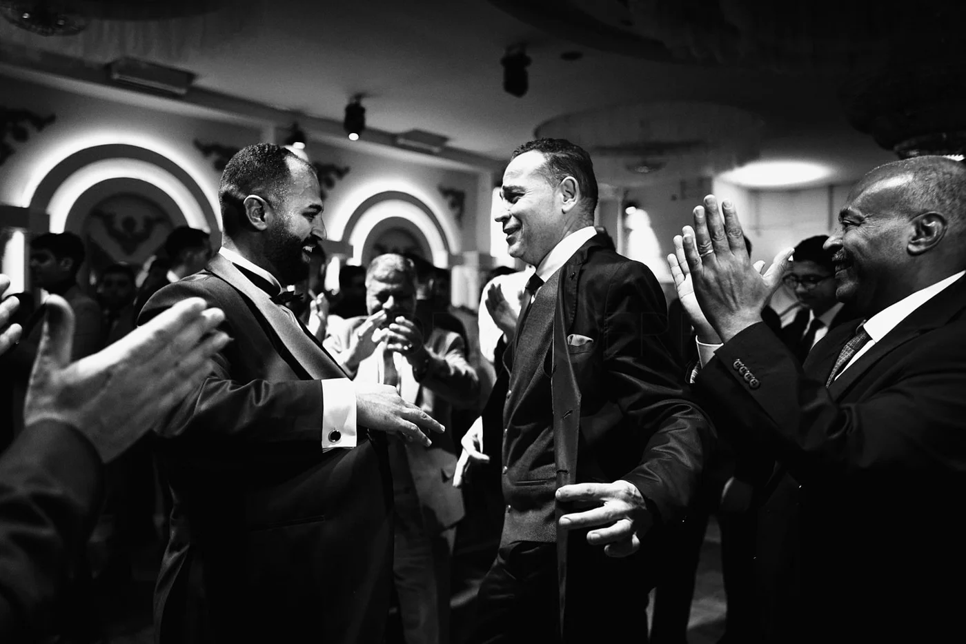 Black and white photo of three men in suits laughing and smiling at each other during a formal celebration or event, with other guests in the background.