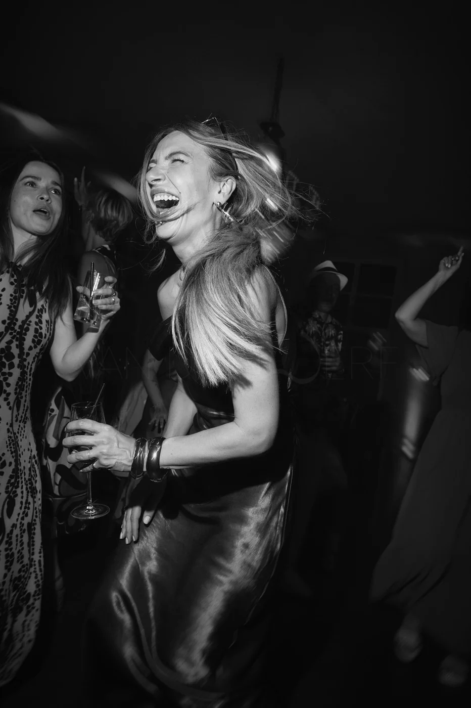 Black and white photo of women dancing and laughing at a party, holding drinks, in a lively atmosphere.