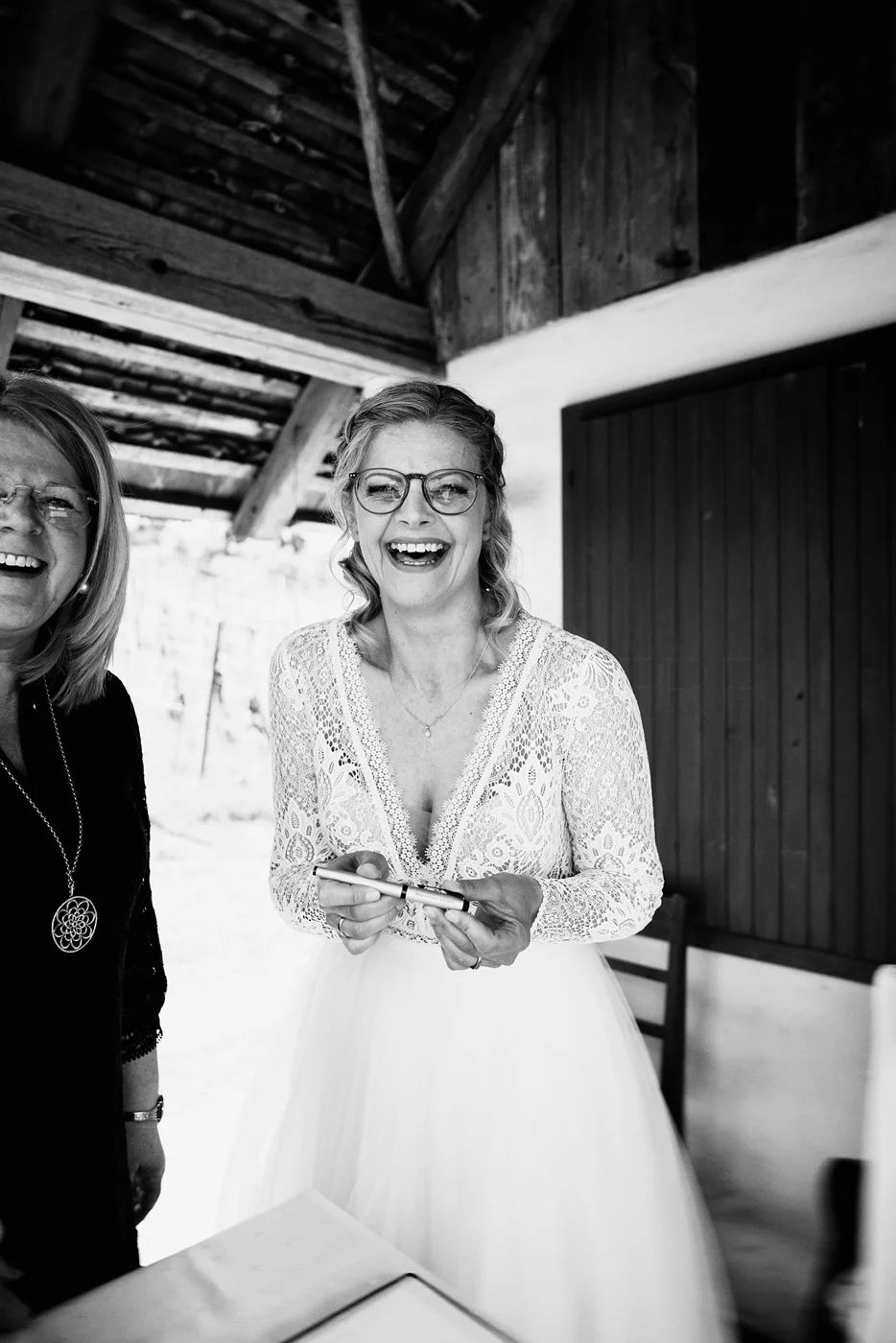 Black and white photo of women smiling and laughing, with one woman in a wedding dress holding a pen.