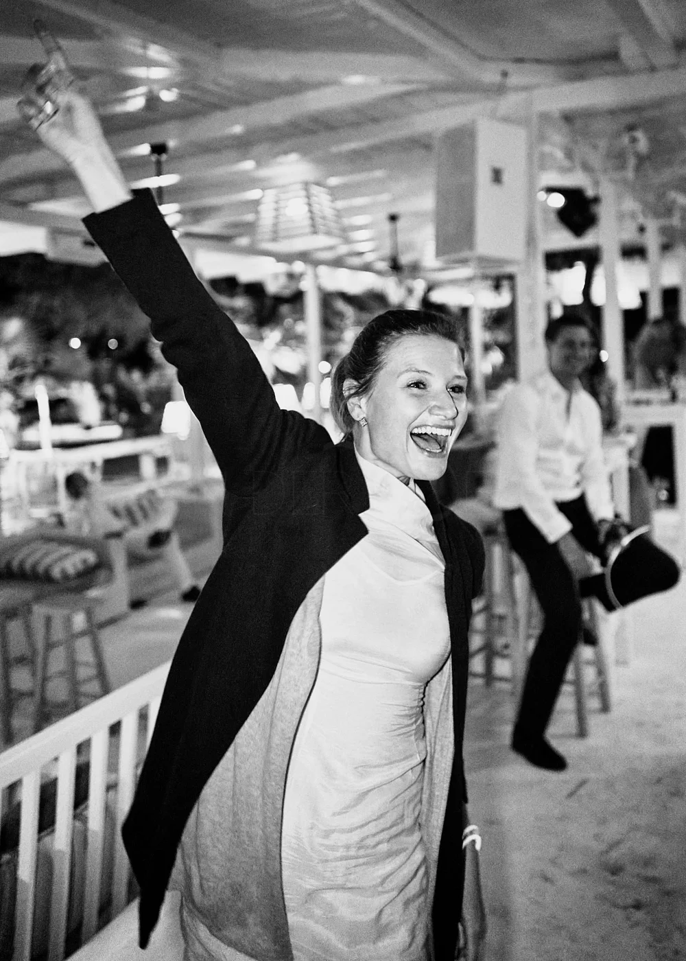 A woman smiling and raising her arm joyfully in a lively indoor setting, with another woman in the background holding a camera.
