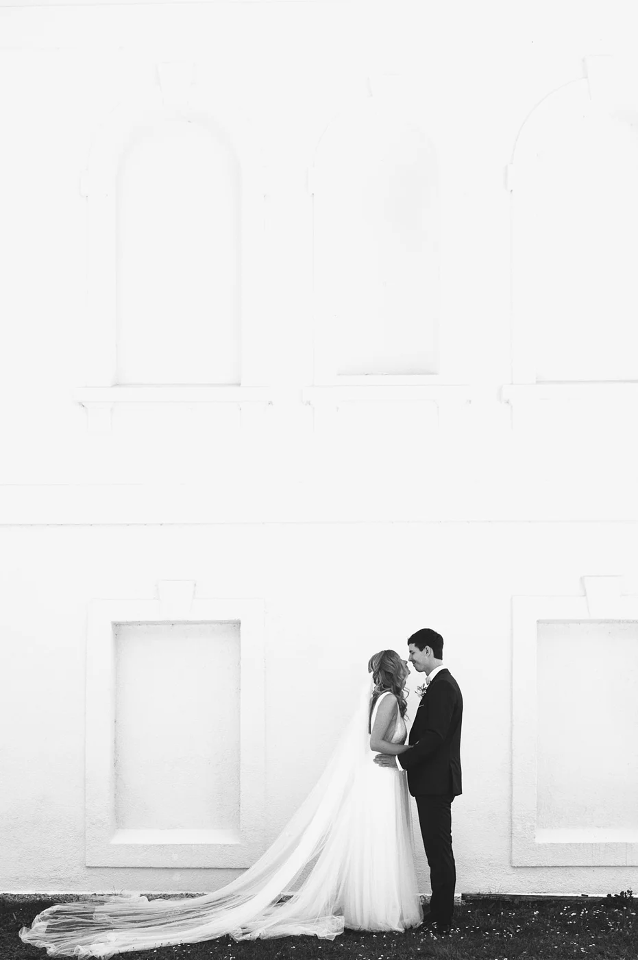 Black and white photo of a bride and groom standing close together, smiling and looking into each other's eyes, in front of a white wall with rectangular and arched windows.