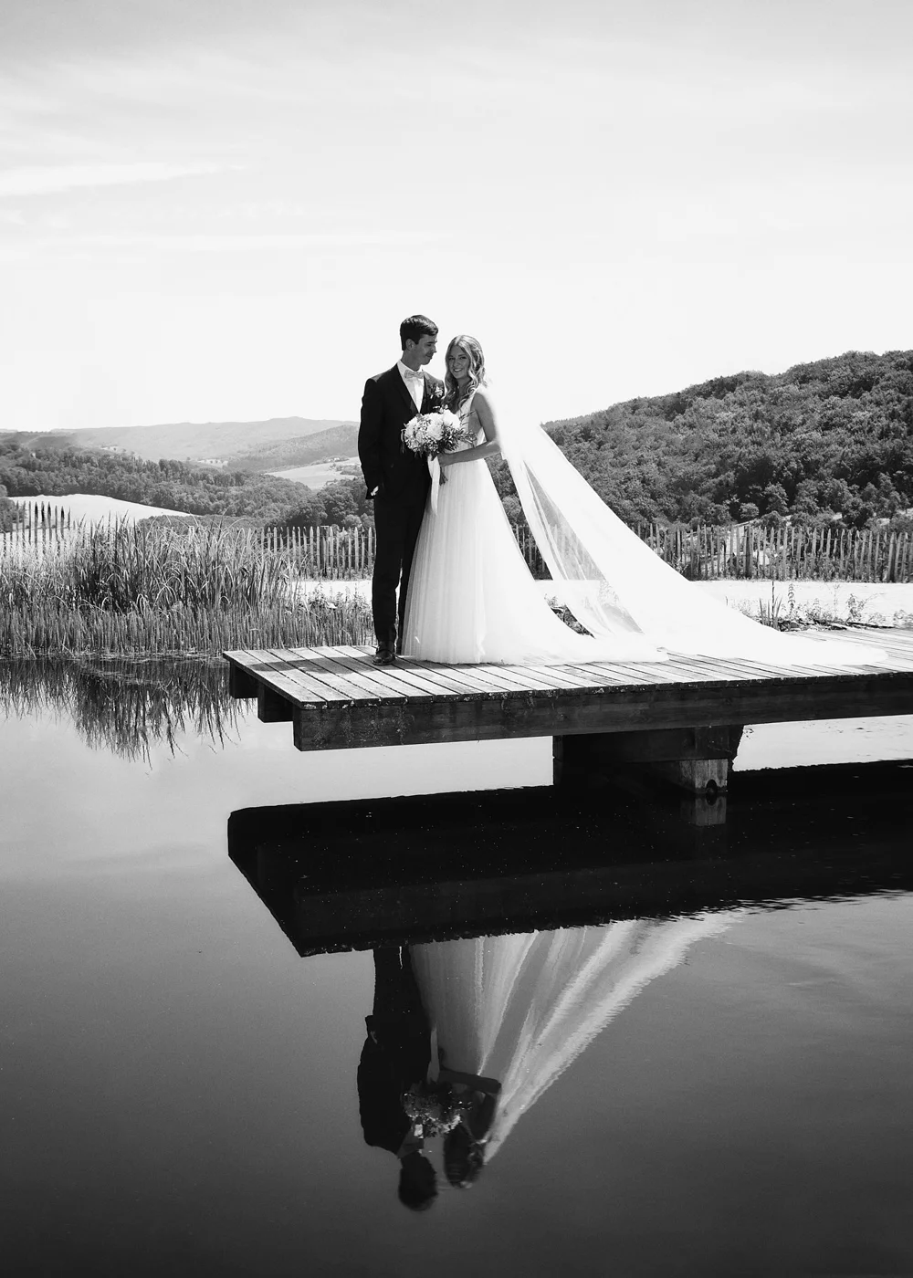 Black and white photo of a bride and groom standing on a wooden dock near water, holding a bouquet, with a scenic landscape of hills in the background, and their reflection visible in the water.