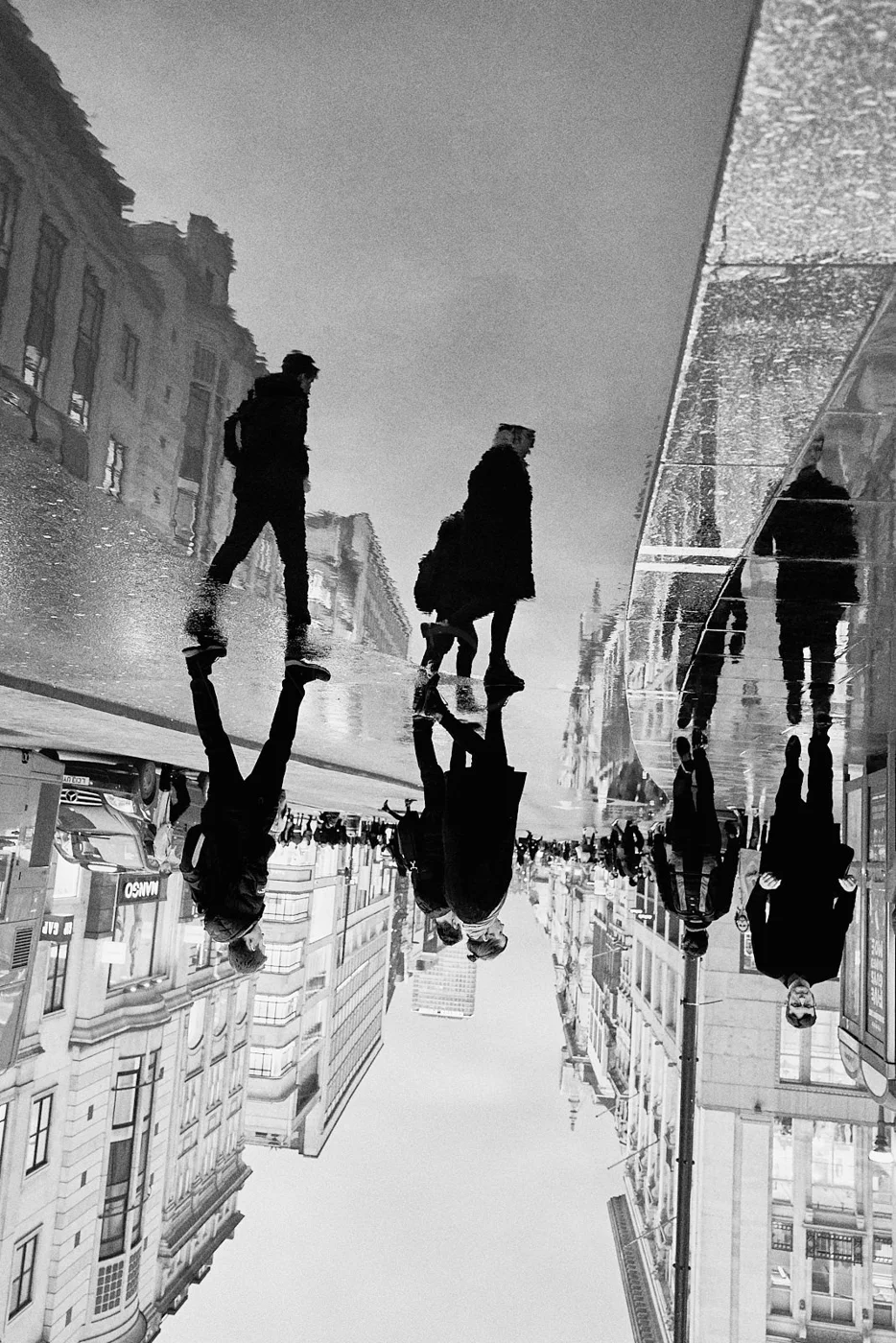 People walking on a city street reflected in a wet pavement, with buildings in the background, in black and white.