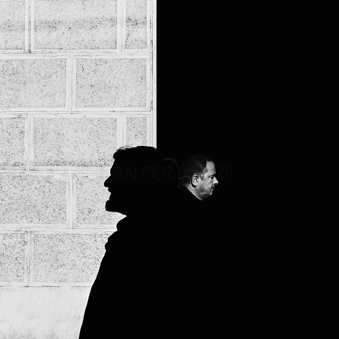 Silhouettes of two men facing opposite directions, split by a vertical line, with one side showing a textured brick wall and the other side in darkness.