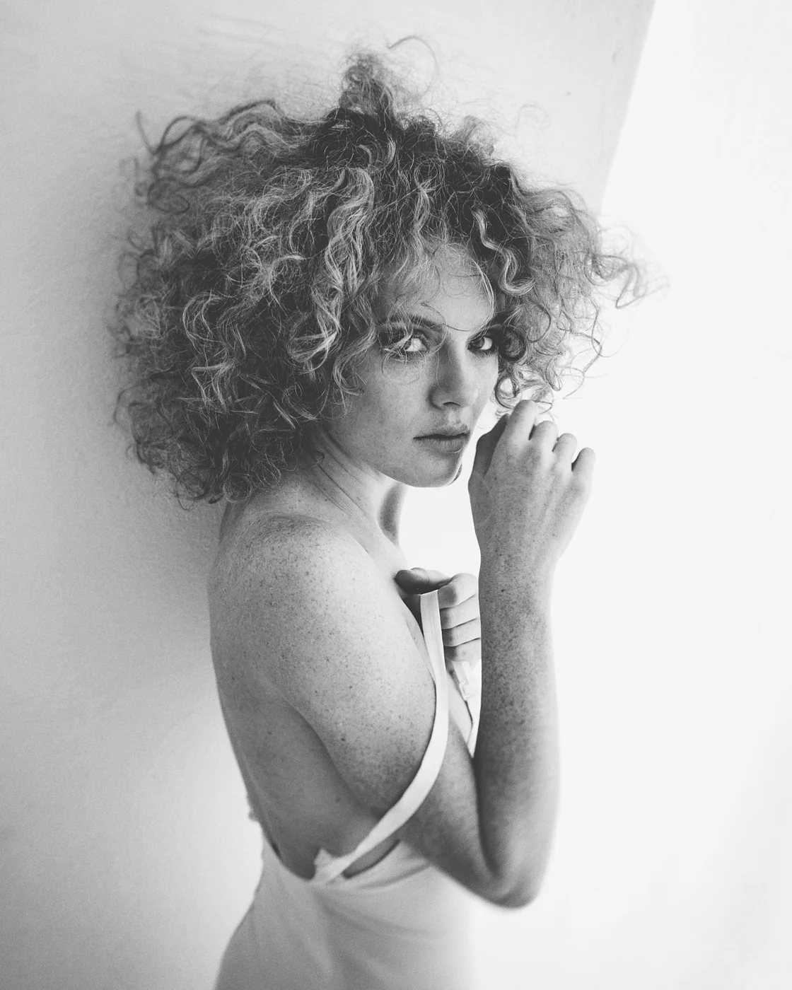 Black and white photo of a woman with curly hair looking at the camera, with one hand near her face, wearing a sleeveless top.