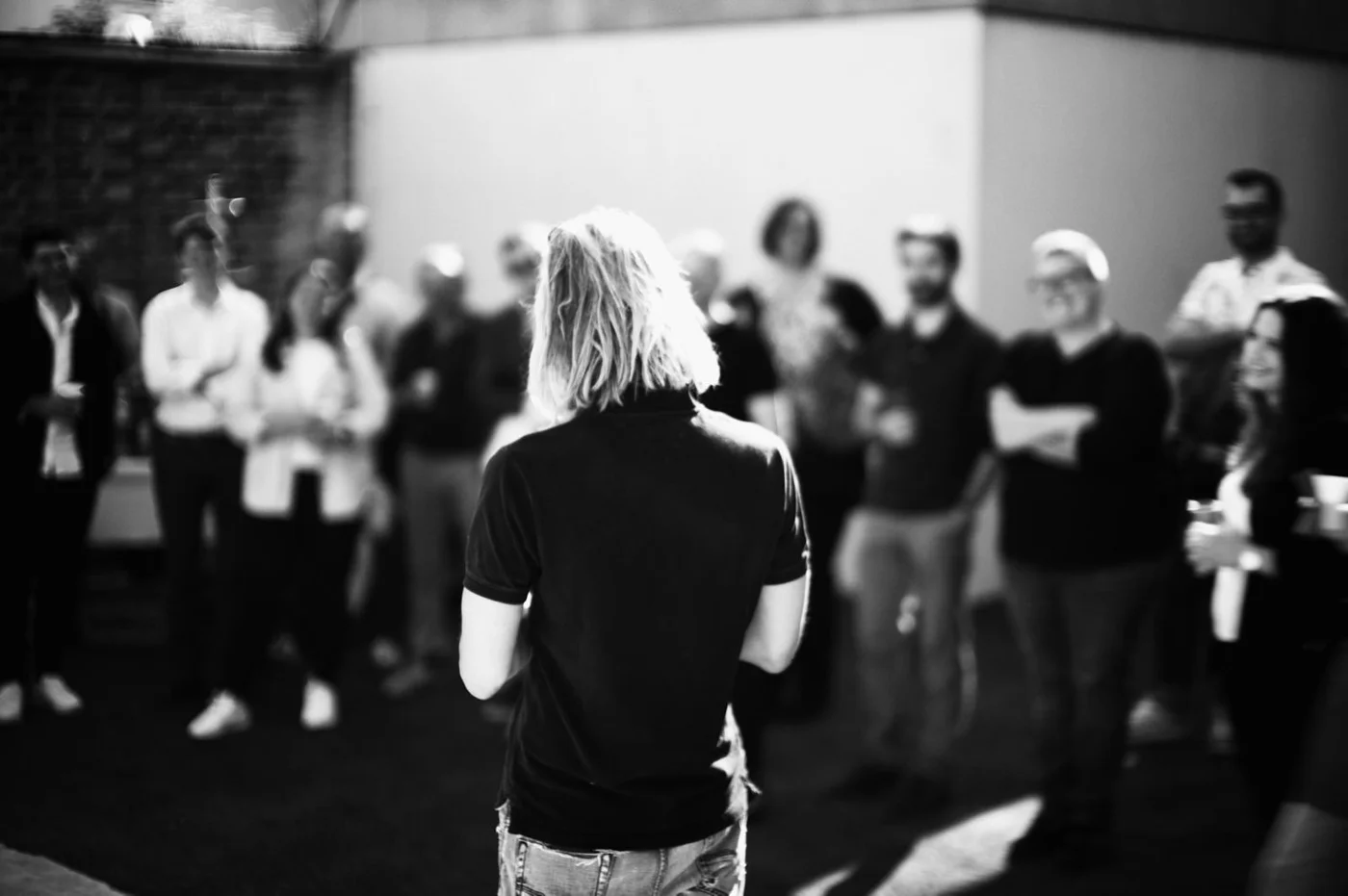 A woman with shoulder-length blonde hair and casual clothing stands with her back to the camera, addressing a group of people in a garage or industrial space, suggesting a presentation or informal meeting.