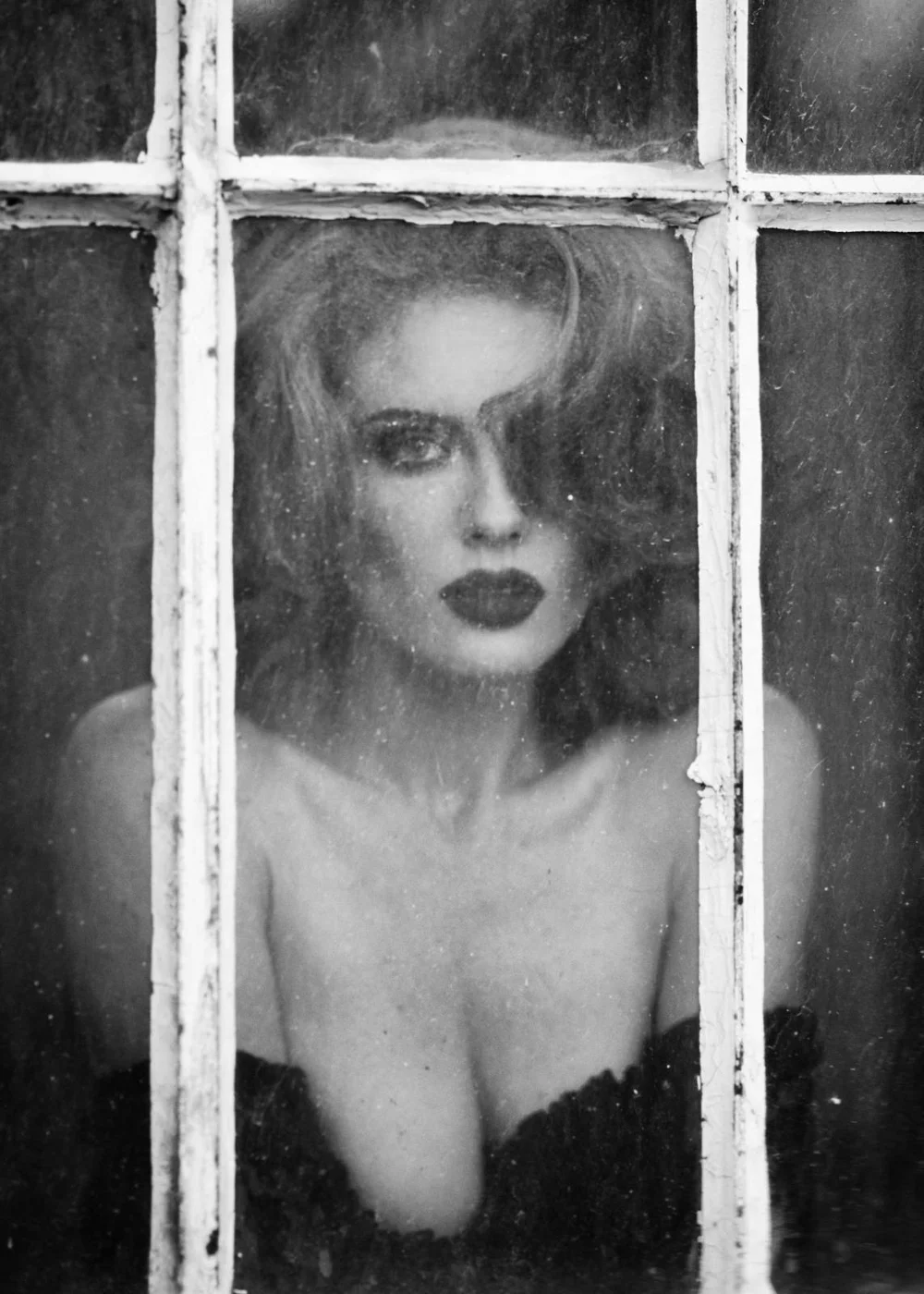 Black and white photo of a person with styled hair and makeup looking through an old window.