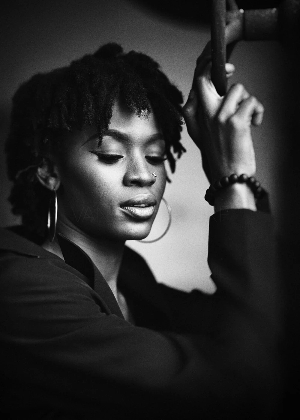 Black and white portrait of a person with natural hair, wearing a blazer, large hoop earrings, and a beaded bracelet, looking down with a thoughtful expression.