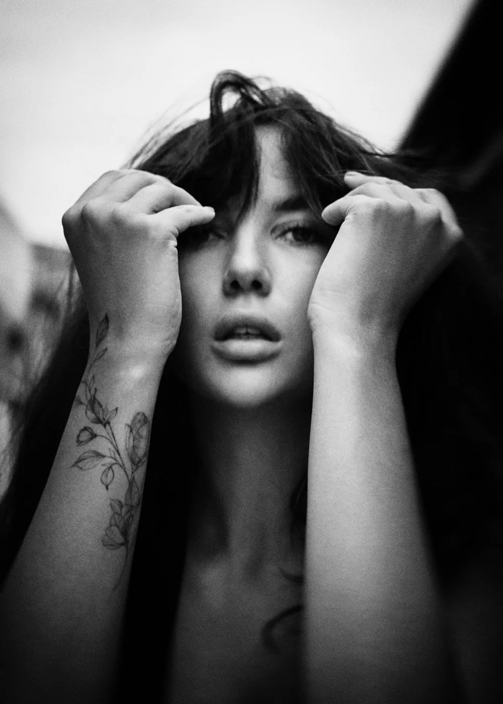 Black and white close-up portrait of a person with long hair. They have their hands raised near their face, and there is a floral tattoo on their forearm.