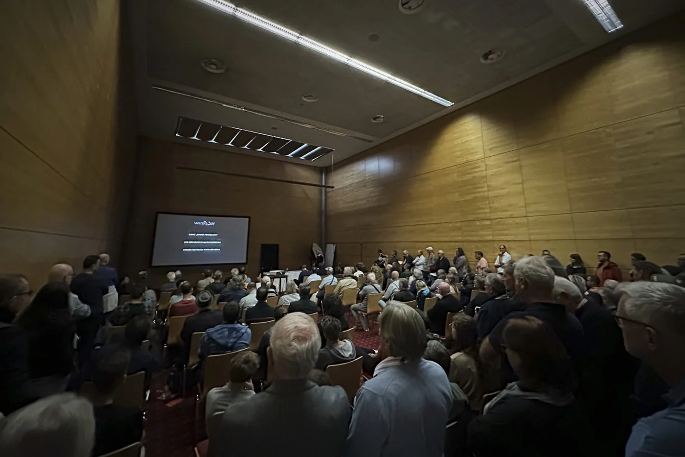 Crowded conference room with audience watching a presentation on a large screen.
