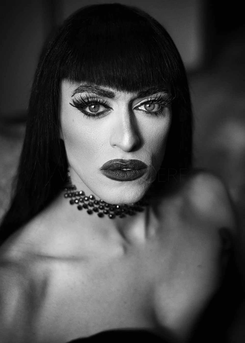 A black and white close-up portrait of a woman with dark straight hair, bold makeup, including dark lipstick and dramatic eye makeup, wearing a beaded choker necklace.