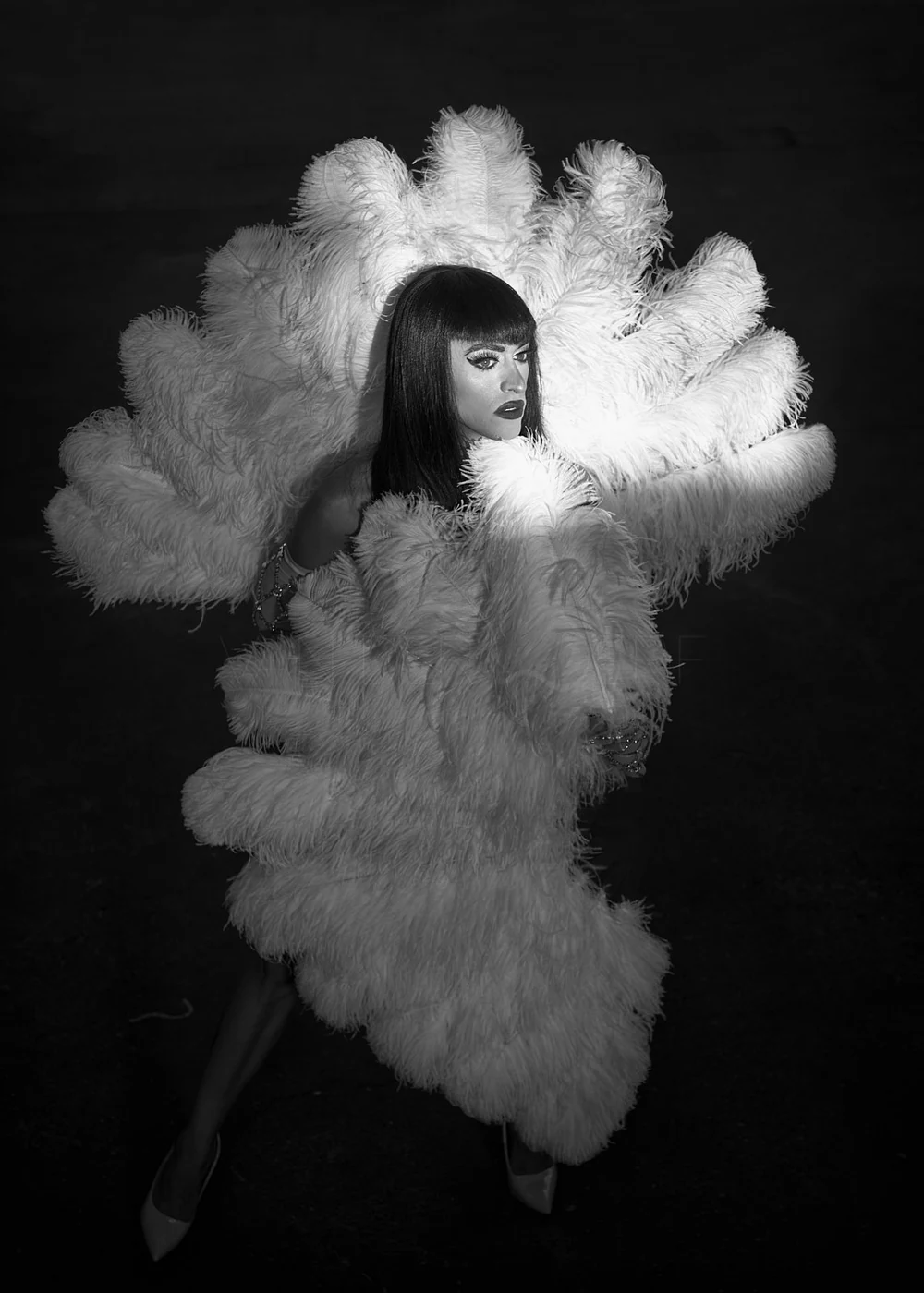 A woman with dark hair and bangs poses with a large, fluffy white feathered costume in a black and white photograph.