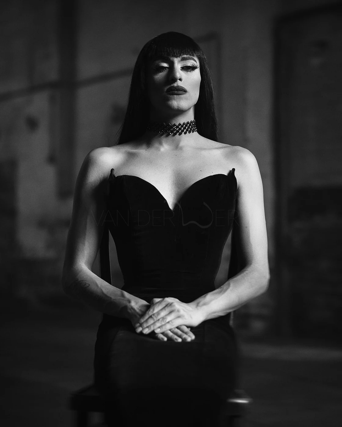 A black and white portrait of a woman with dark hair and bangs, wearing a strapless black dress and a choker, standing with her hands clasped in front of her, in a dimly lit room.