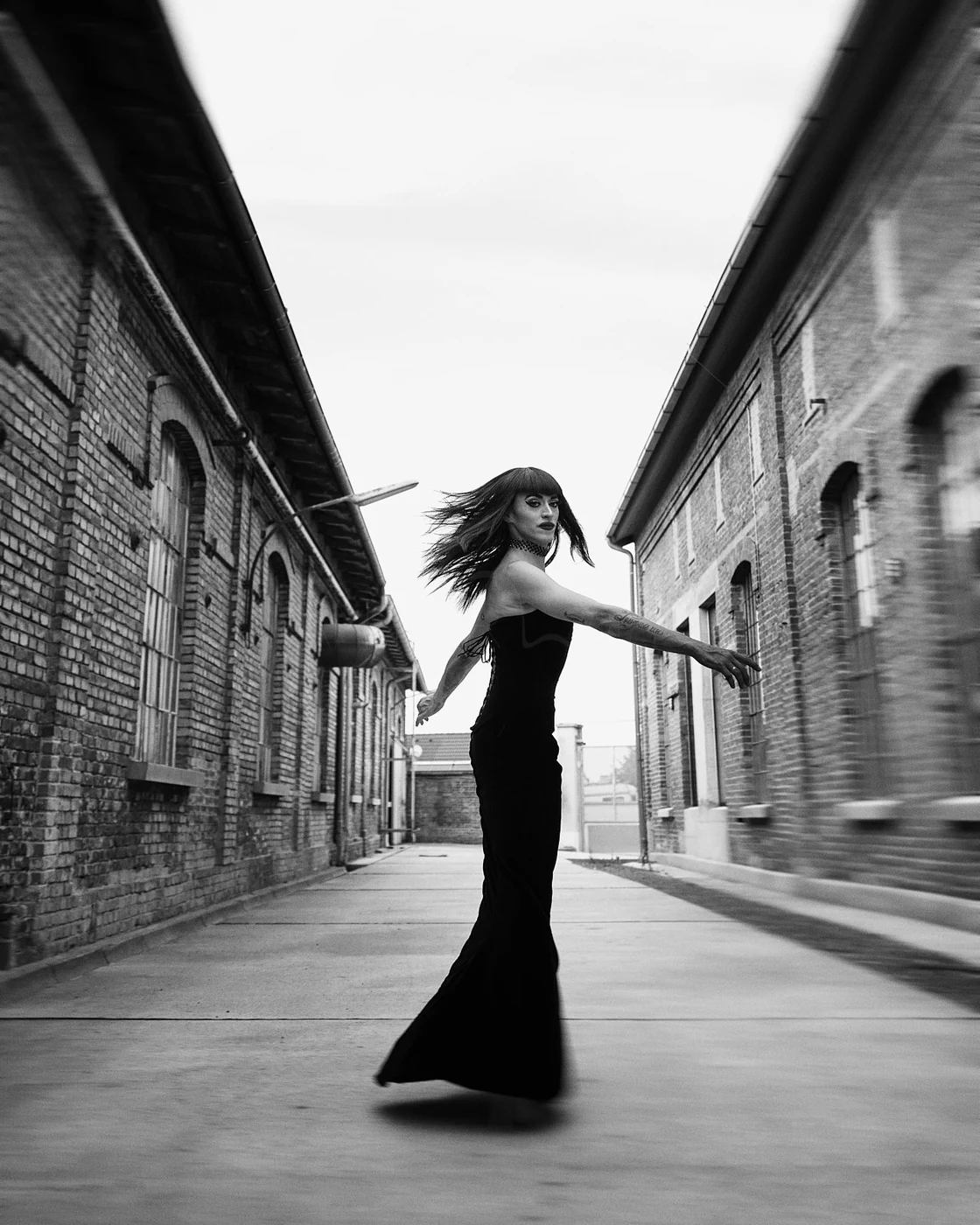 Black and white photo of a woman in a long dress with tattoos, standing between two brick buildings, looking over her shoulder with hair blowing, in an alleyway.