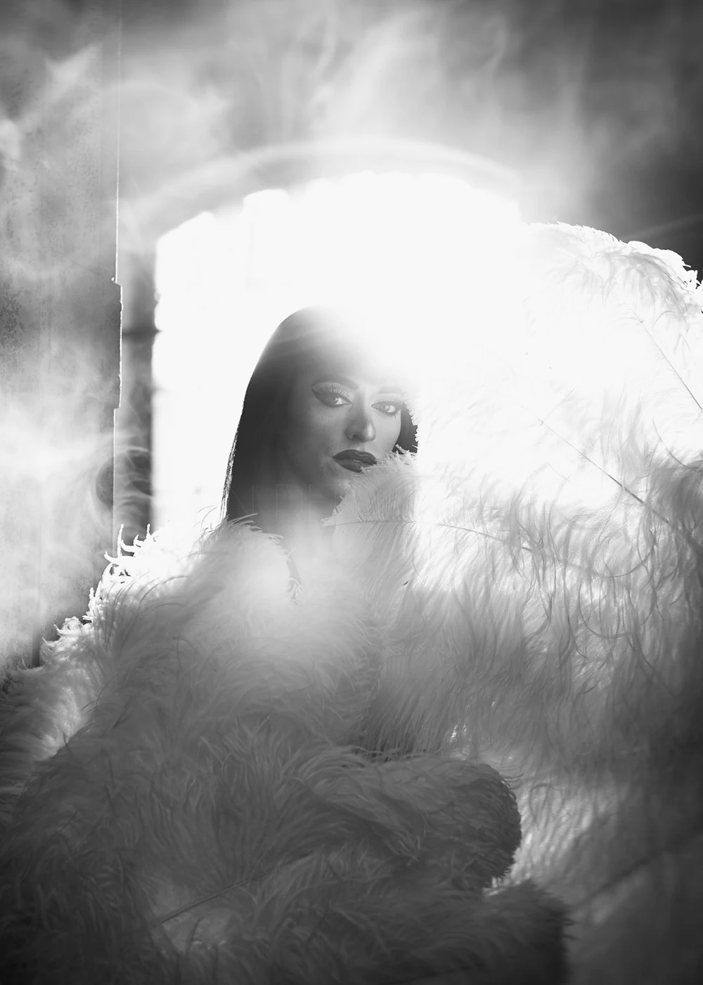 A woman with makeup and dark hair, surrounded by large, fluffy feathers, looking into the camera with backlighting creating a halo effect.