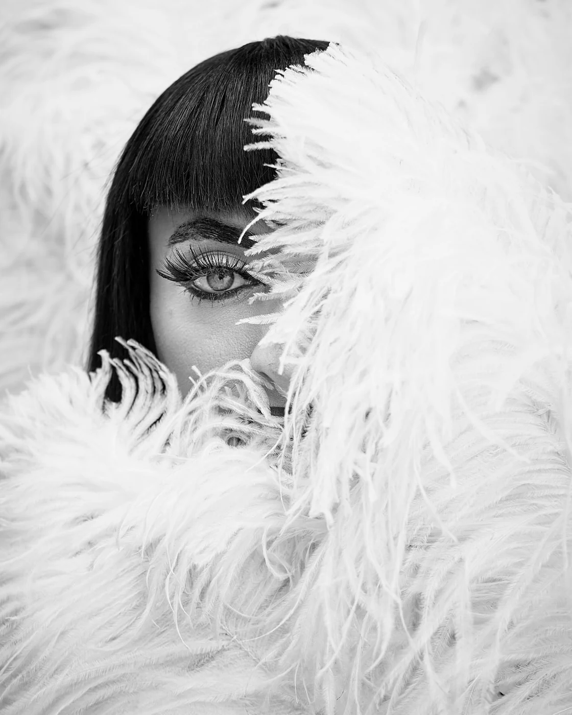 A woman with dark, straight hair and bangs, gazing through white feathers that partially cover her face and eye.