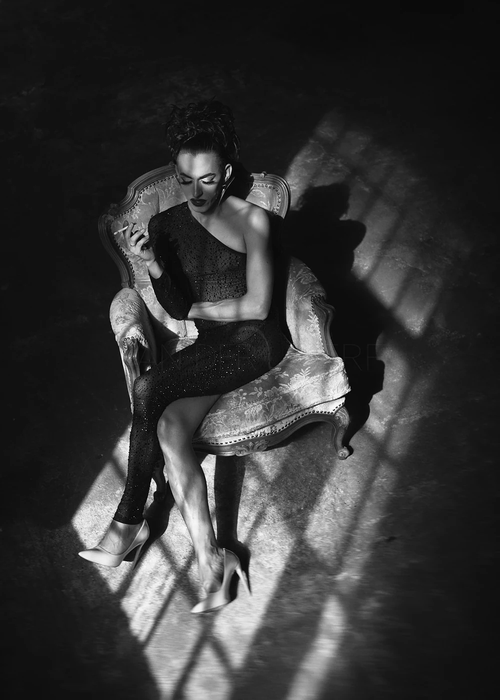 A woman sitting on an ornate vintage chair, wearing a black off-shoulder dress and high heels, smoking a cigarette in a dimly lit room with strong shadows.