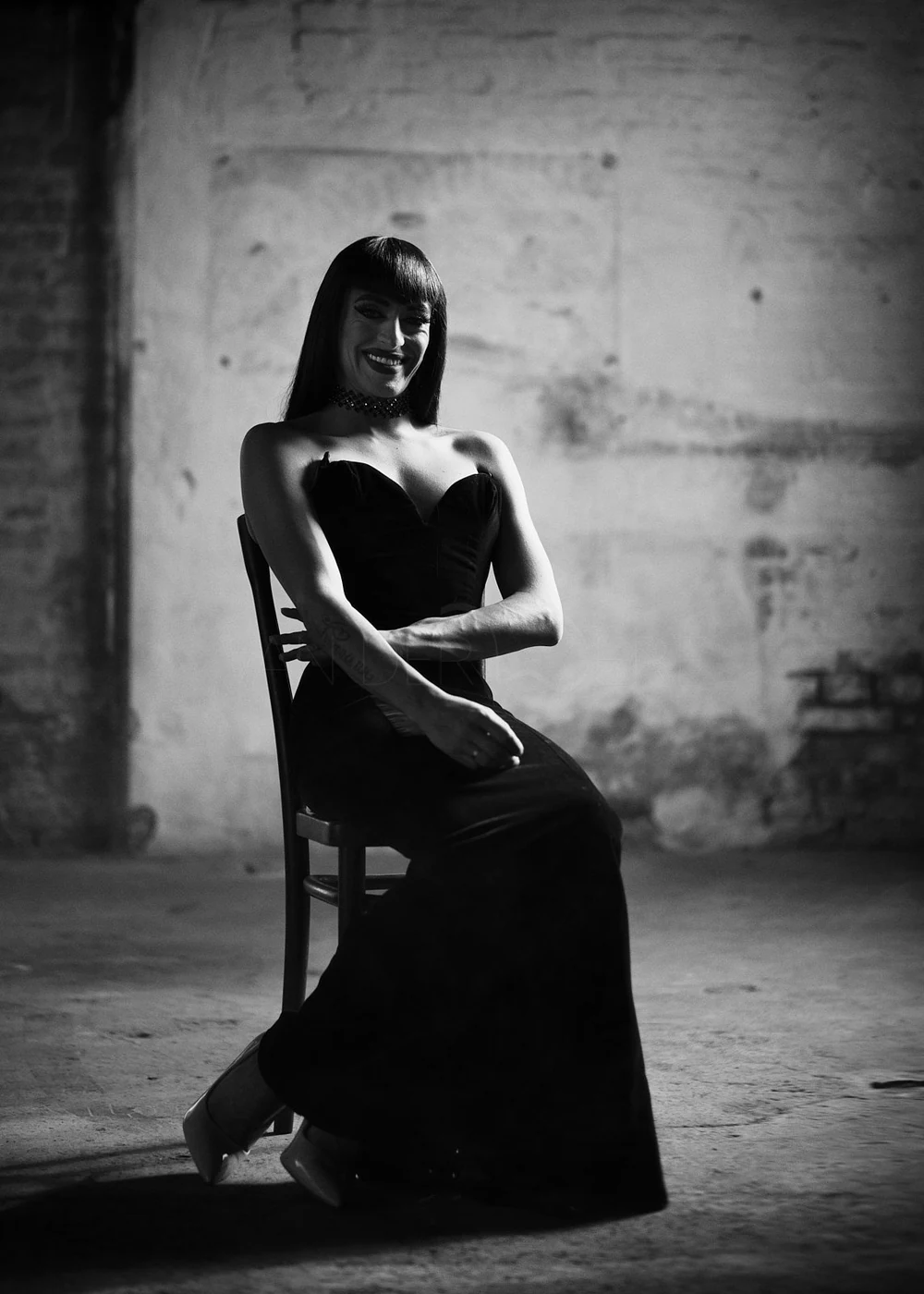A woman in a strapless black dress sitting on a chair with her arms crossed, smiling, in a dimly lit room with a textured wall background.
