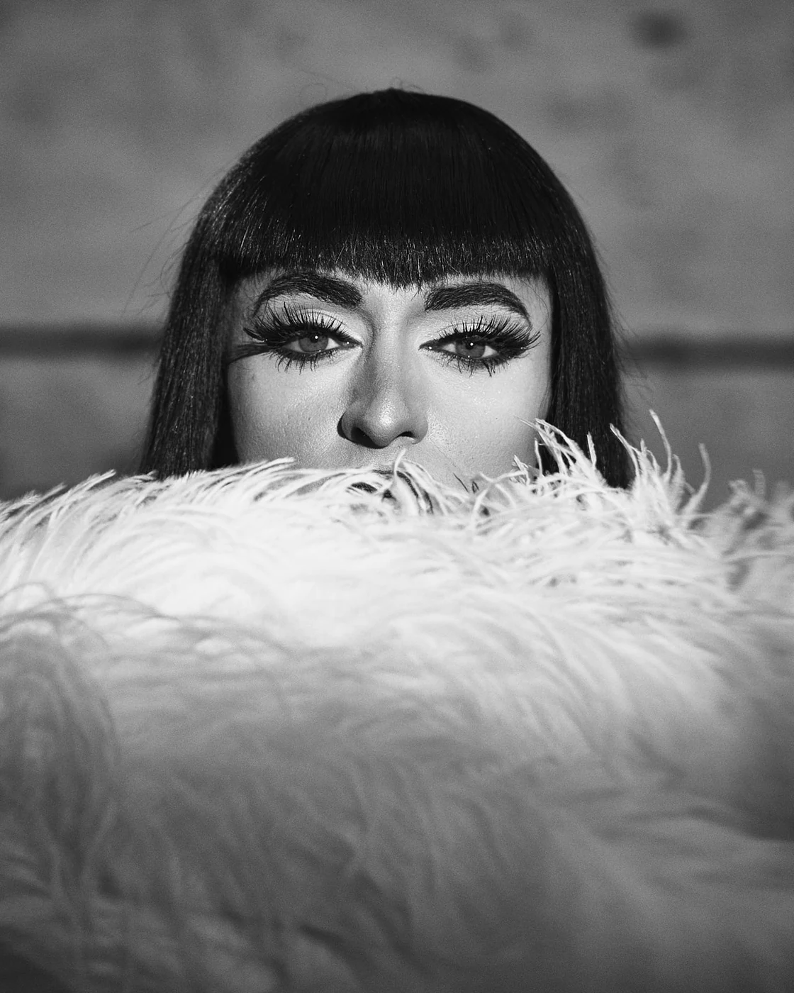 Close-up black & white photo of a woman with dark straight hair and bold eye makeup, partially hidden behind fluffy white feathers.