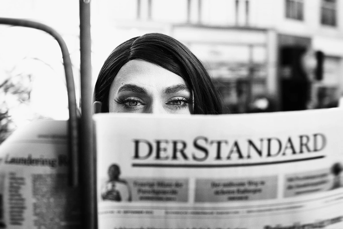 A woman with makeup and dark hair reading a newspaper titled 'DER STANDARD' outdoors.