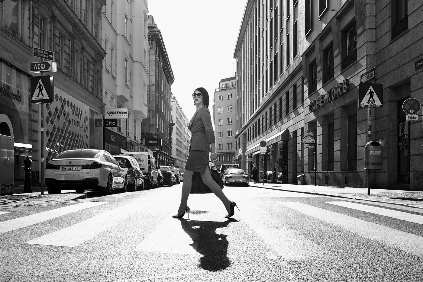 Black and white photo of a woman crossing a city street at a crosswalk. She is dressed in a suit, wearing high heels, and carrying a handbag. Tall buildings and parked cars line the street.
